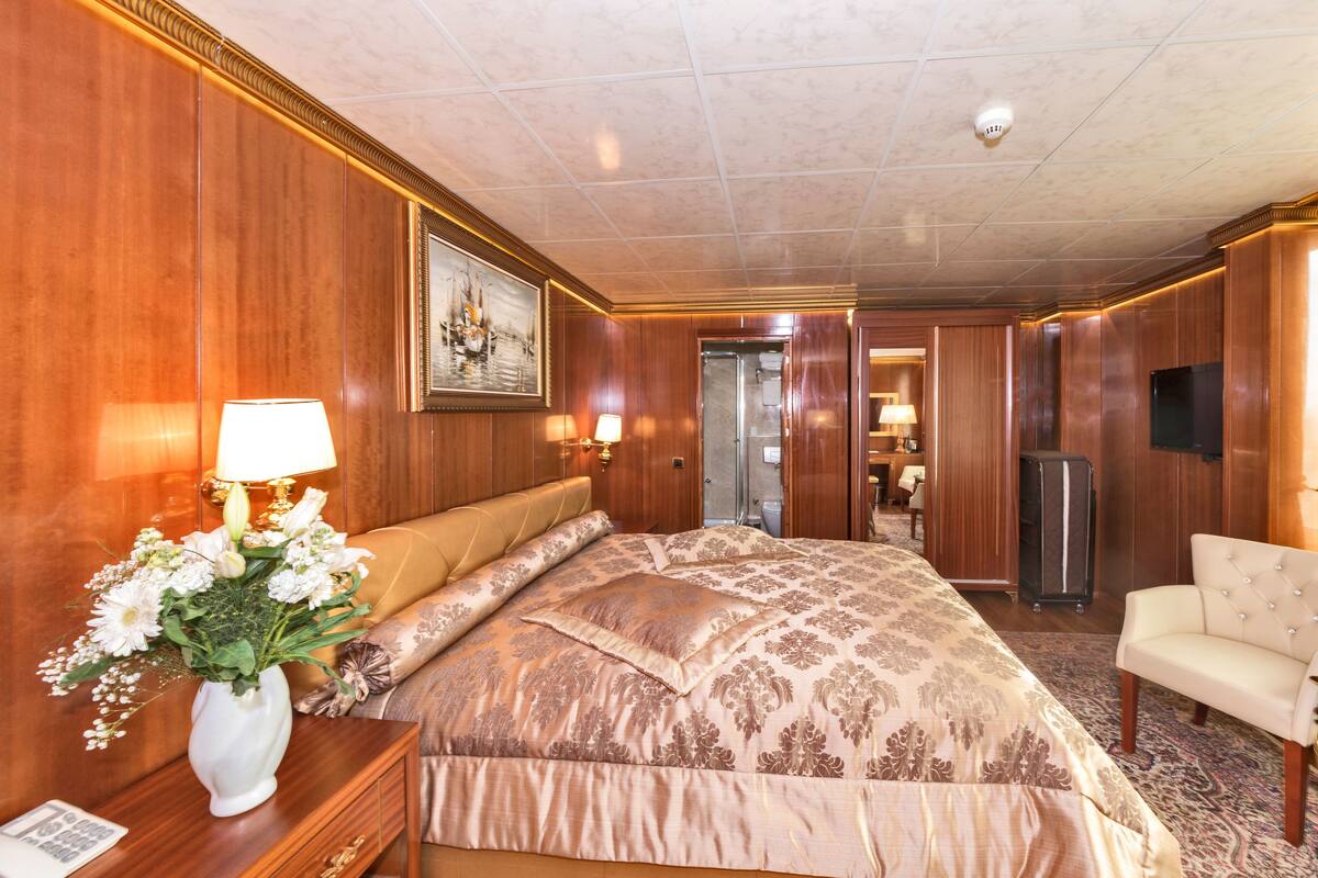 A hotel room with a large bed, a sitting area with a chair, a desk with a lamp, a vase of flowers, and a painting on the wall.