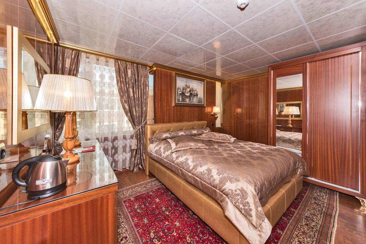 A hotel room with a bed, a desk with a kettle, a mirror, and a red carpet.