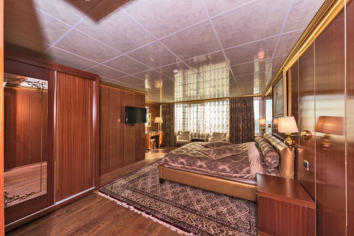 A spacious bedroom with a large bed, a wooden wardrobe, a television, and a window with curtains.