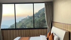 Una habitación de hotel moderna con una cama grande, cabecera de madera y vistas a un paisaje montañoso a través de ventanas de gran altura.