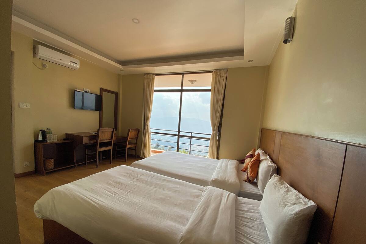 A hotel room with two beds, a desk with chairs, a TV, and a large window with a view.
