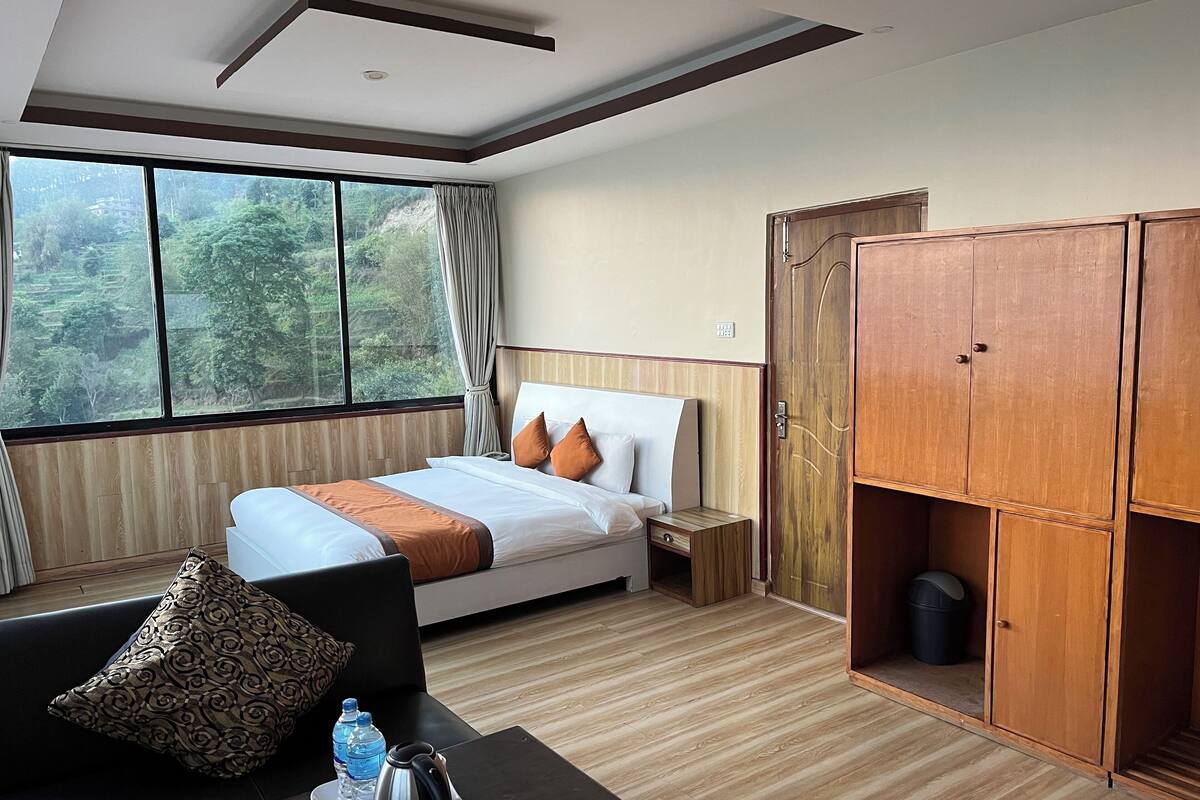 A hotel room with a bed, a wooden wardrobe, a small table with a water bottle and a kettle, and a view of greenery through large windows.