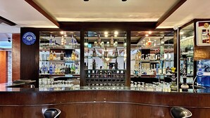 Una zona de bar con dos taburetes negros, estanterías llenas de botellas y vasos, y un techo con vigas a la vista y luces colgantes.
