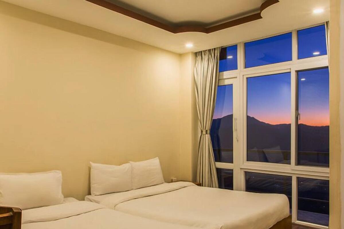 A double bed with a wooden headboard, a nightstand, and a window with a view of a sunset over mountains.