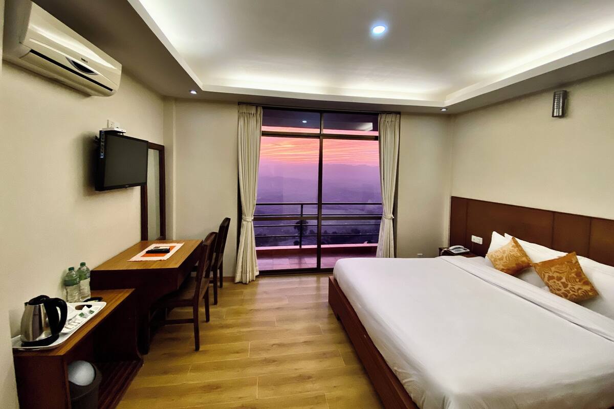 A hotel room with a bed, a desk with a chair, a television, and a view of a sunset through the window.