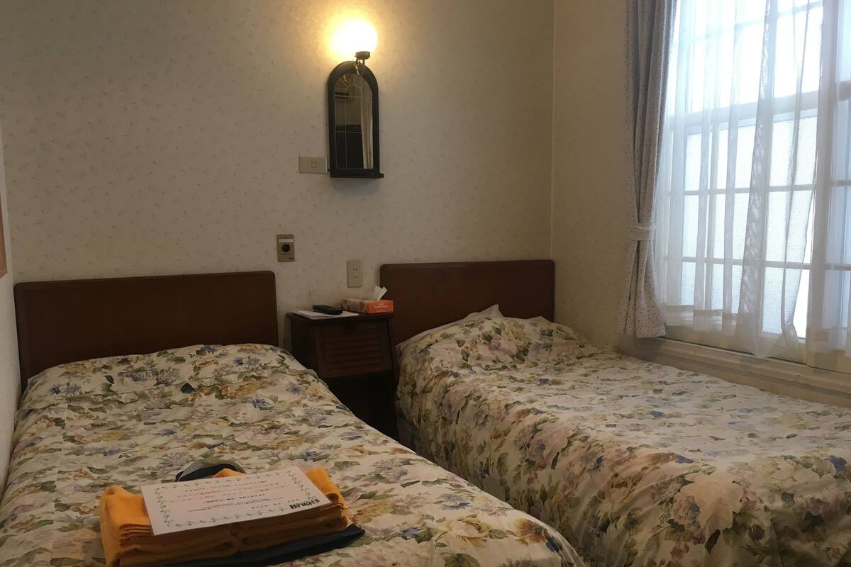 A hotel room with two beds, floral bedding, a nightstand with a lamp, and a window with sheer curtains.