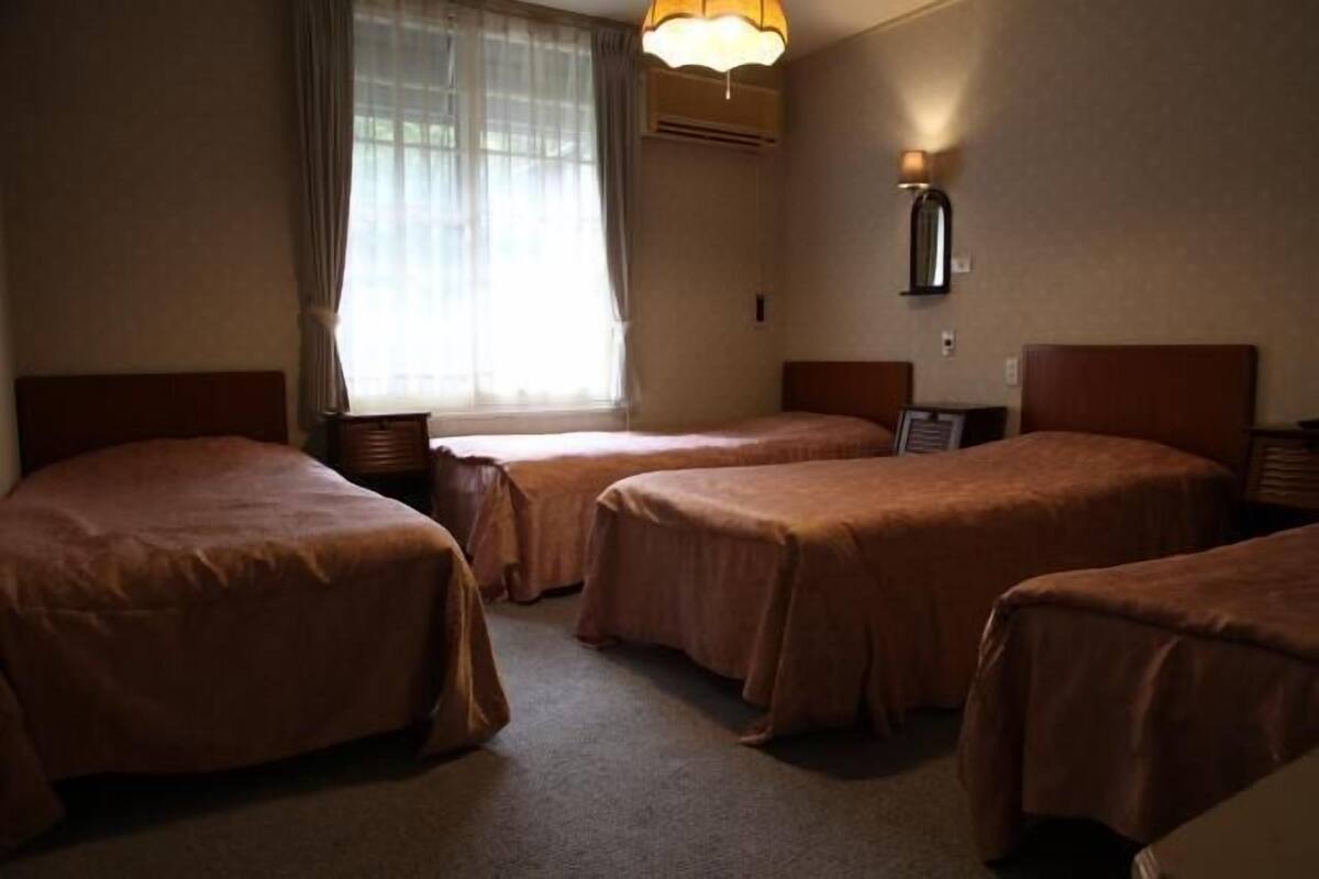 A hotel room with three single beds, a window with sheer curtains, an air conditioning unit, and a wall-mounted lamp.