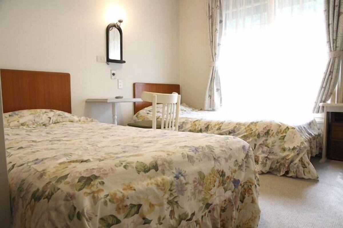 A hotel room with two beds, a small table, a chair, and a window with curtains.