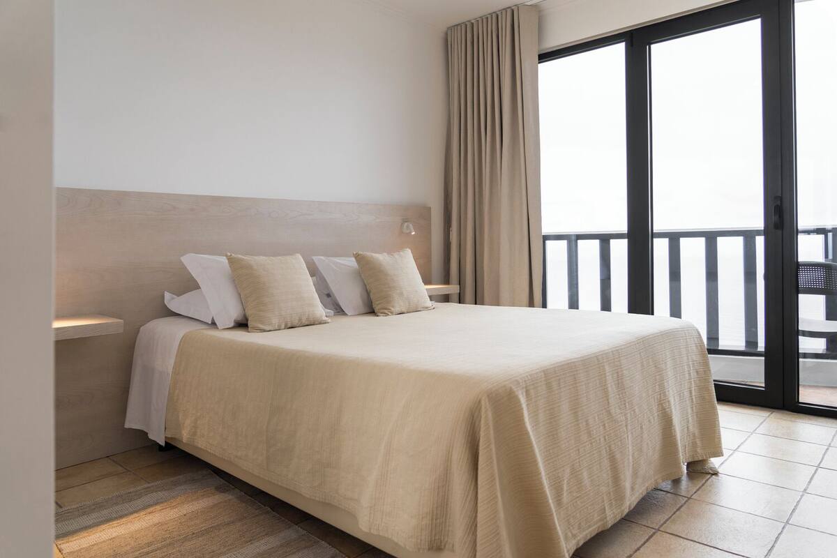 A hotel room with a large bed, a wooden headboard, a balcony with a view, and a sliding glass door.