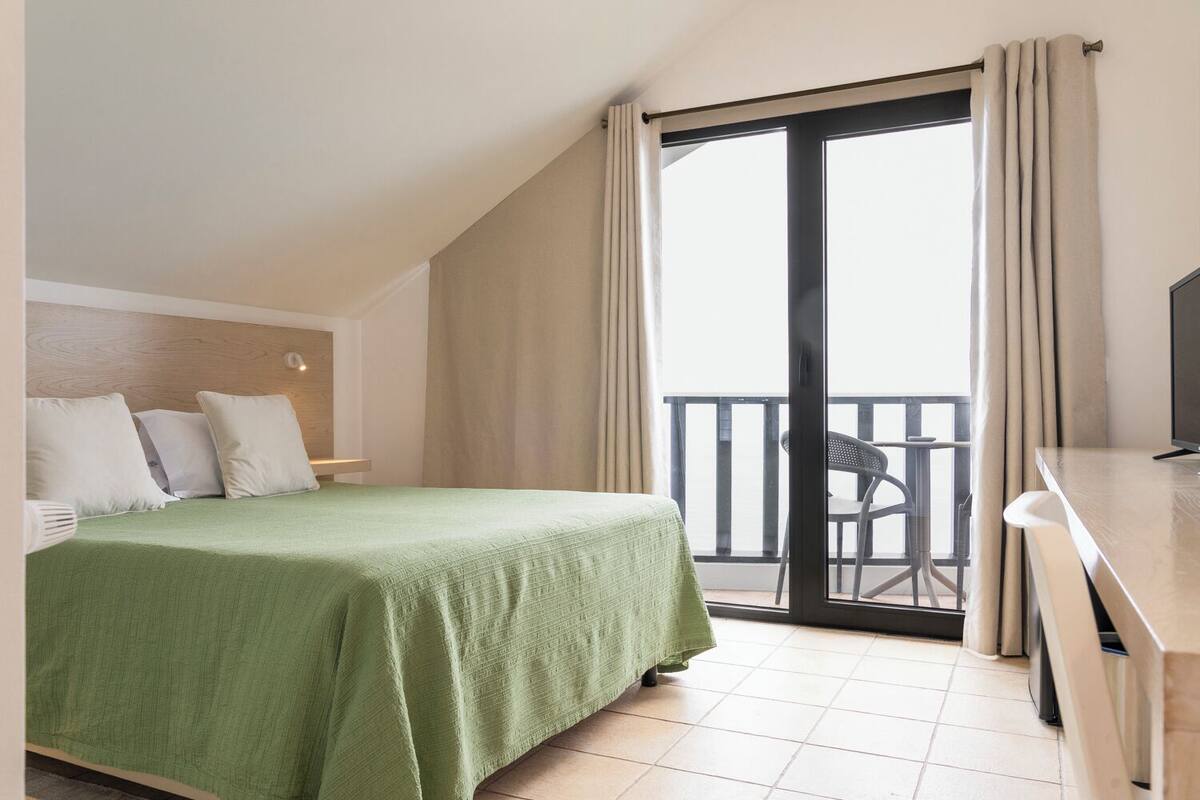 A hotel room with a bed, a desk, a chair, and a balcony with a view.