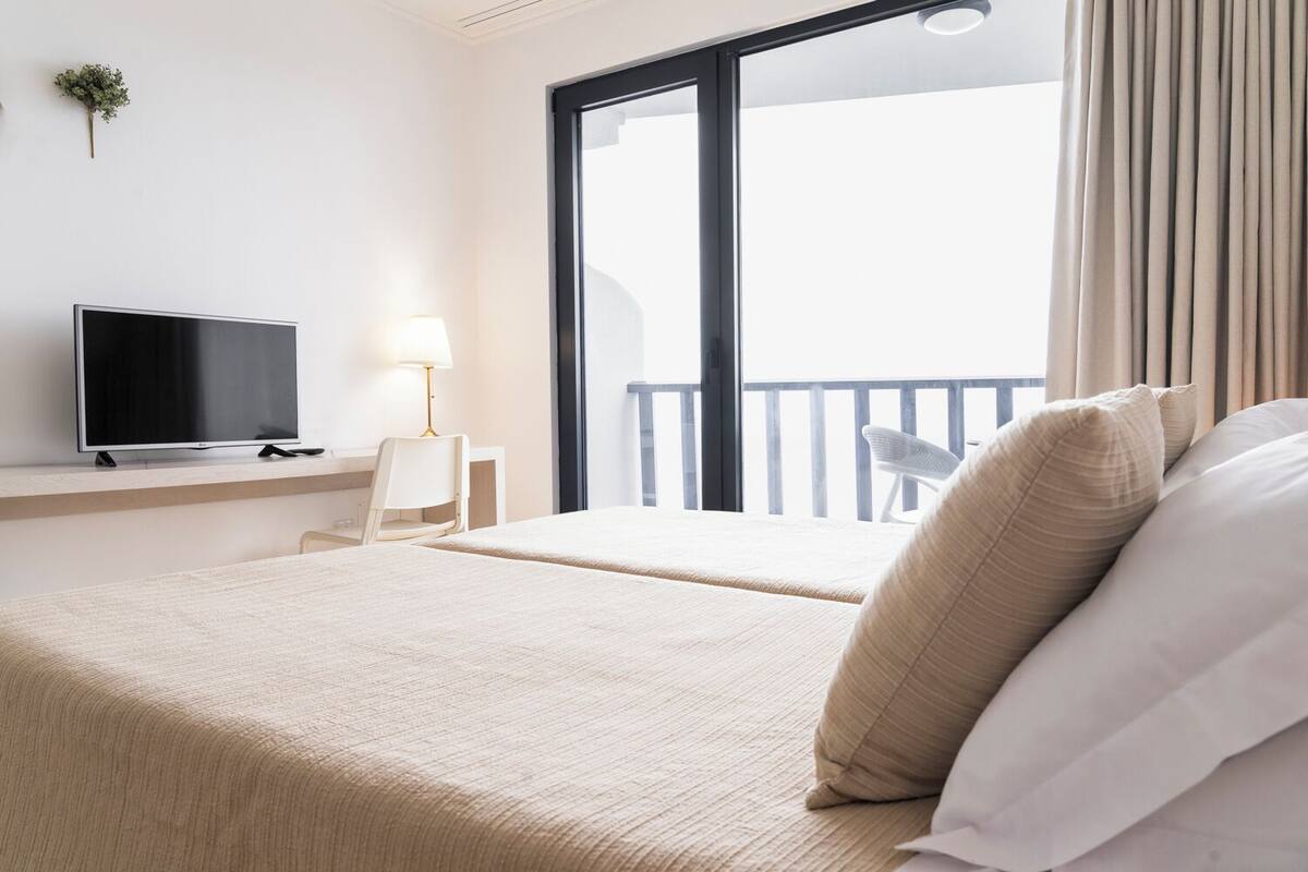 A hotel room with a bed, a TV on a stand, a desk with a chair, and a balcony with curtains.