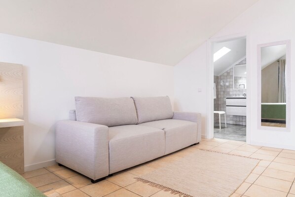Una sala de estar pequeña y moderna con un sofá gris, piso de madera y vista a un baño por una puerta abierta.