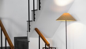 A staircase with black railings and wooden handrails, a standing lamp, and a wicker chair with a white cushion. - Casa das Proteas (Santana)