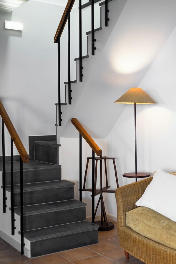 A staircase with black railings and wooden handrails, a standing lamp, and a wicker chair with a white cushion. - Casa das Proteas (Santana)
