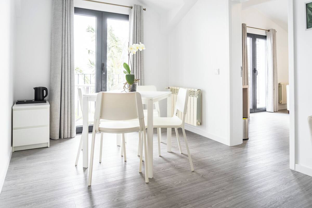 A modern, well-lit room with a dining table and chairs, a radiator, and a view of the outdoors through large windows.