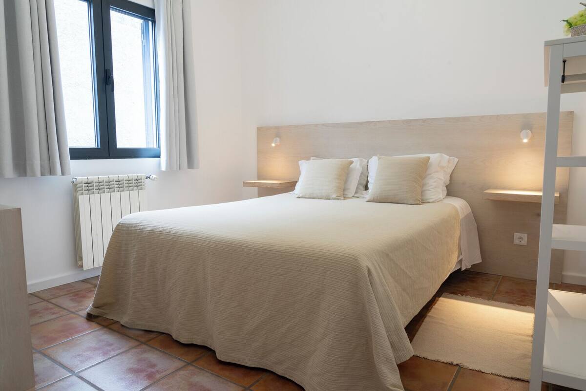 A bedroom with a bed, a radiator, a window with curtains, and a nightstand with a lamp.