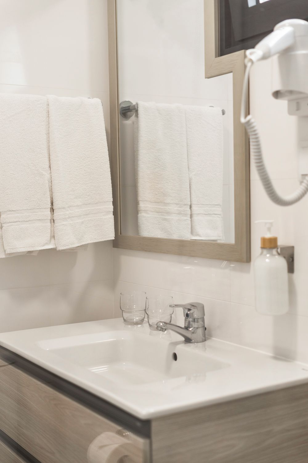 Un baño con un lavamanos blanco, dos toallas colgadas en la pared, un espejo y un secador de pelo sobre el mostrador.