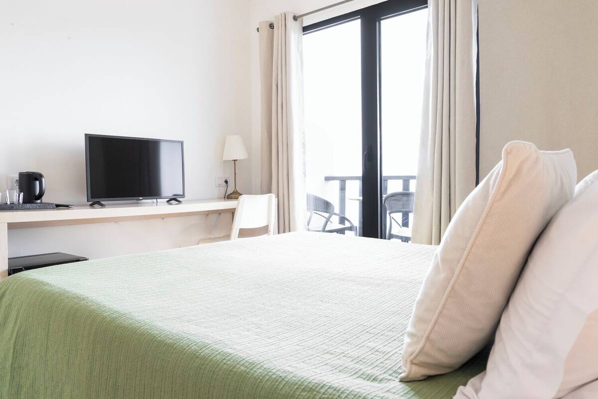 A hotel room with a bed, a desk with a television, a lamp, and a view of the outside balcony.