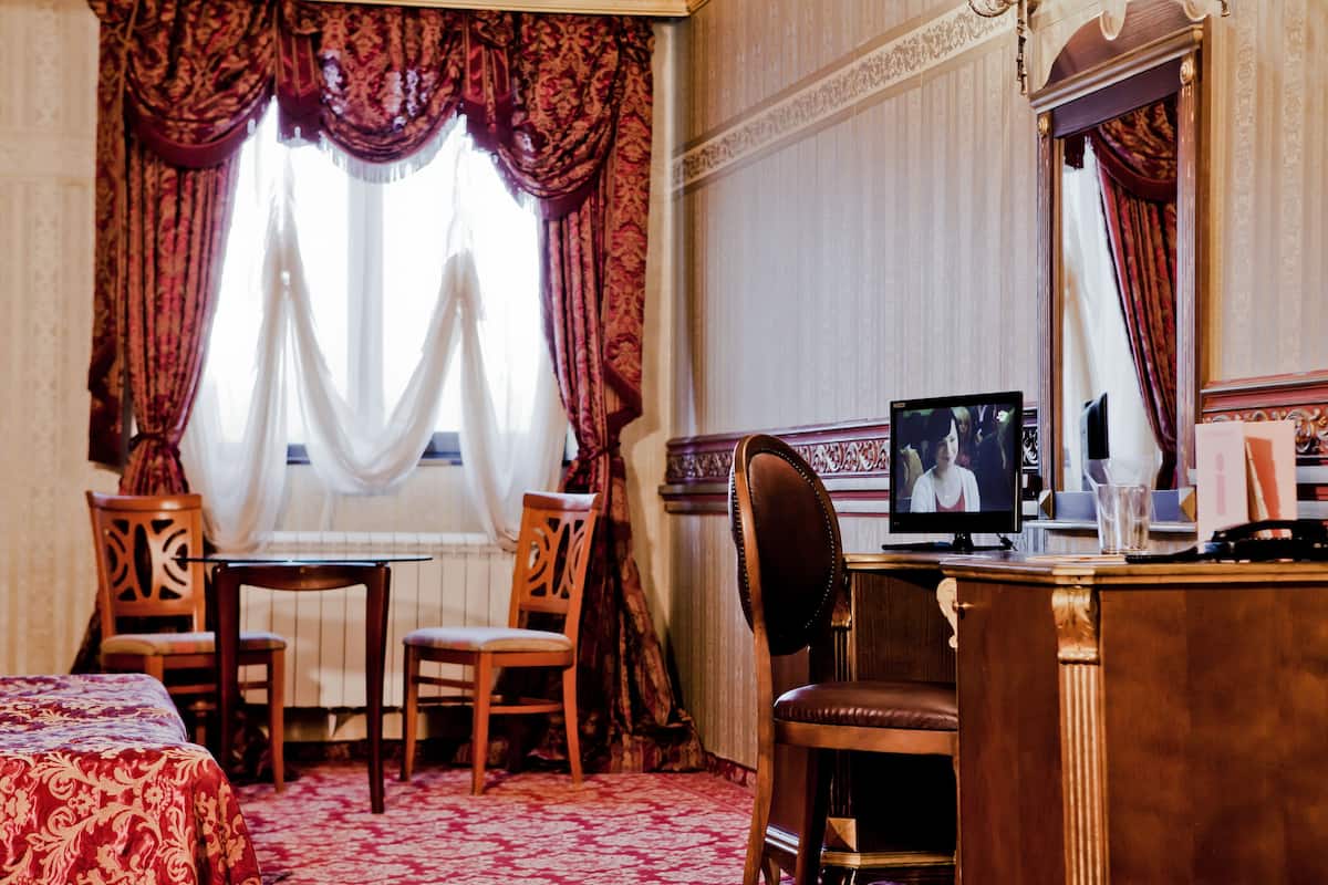 A hotel room with a bed, a desk with a computer, two chairs, and a large window with curtains.