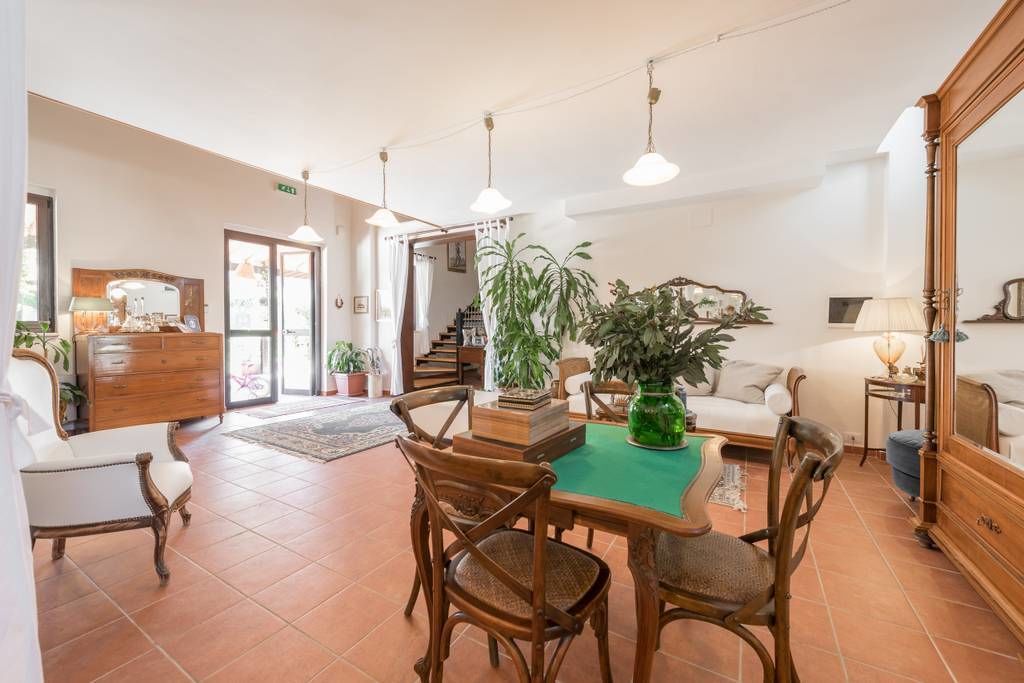 A spacious living area with a dining table, chairs, and a sofa. There are multiple potted plants and a large mirror.