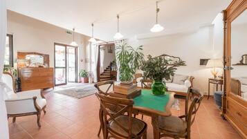 A spacious living area with a dining table, chairs, and a sofa. There are multiple potted plants and a large mirror.