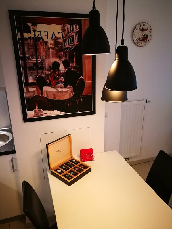 A dining area with a table, chairs, a framed picture, a clock, and a box of chocolates.