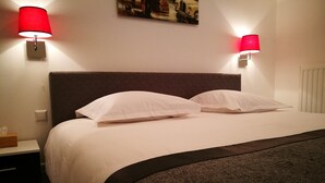 Una cama bien hecha con sábanas blancas y dos almohadas, flanqueada por dos lámparas de pared con abat-jour rojos, un cuadro enmarcado encima de la cama y un radiador blanco en la pared.