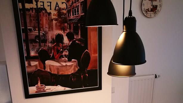 Una obra de arte en marco que muestra una escena de café, una lámpara colgante negra y un reloj antiguo en la pared.