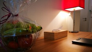 Una mesa de madera con una cesta de plástico transparente que contiene manzanas verdes y rojas, una caja de pañuelos y un dispositivo electrónico negro.