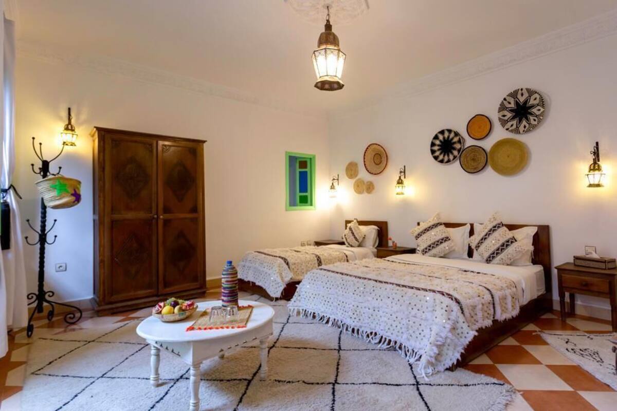 A bedroom with a large bed, two bedside tables, a small round table with a fruit bowl, and a decorative wall hanging.