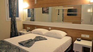 Habitación de hotel con una cama grande, una mesita de noche con teléfono, un espejo sobre la cama y una ventana con cortinas.
