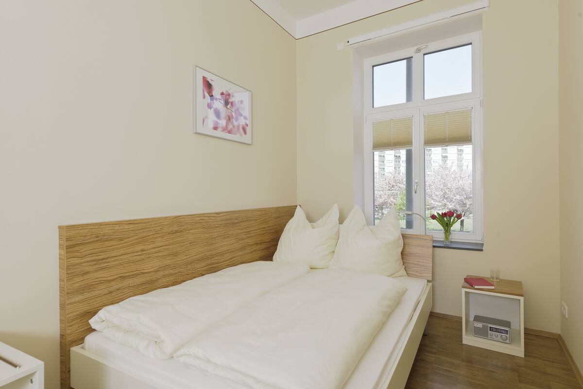 A bedroom with a wooden headboard, white bedding, a bedside table with a radio, and a window with a view of trees and buildings.