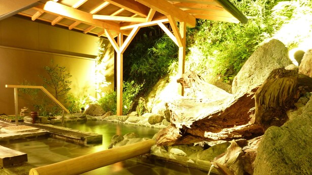 Un onsen japonés tradicional con estructura de madera, un baño de aguas termales y rocas y plantas circundantes.