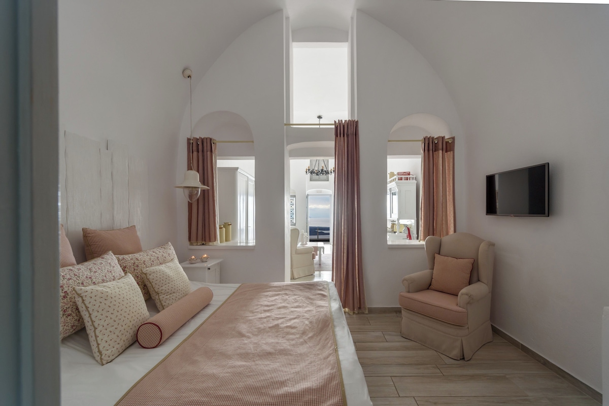 Un dormitorio con una cama grande, una silla, un televisor y vistas al mar a través de un ventanal arqueado.
