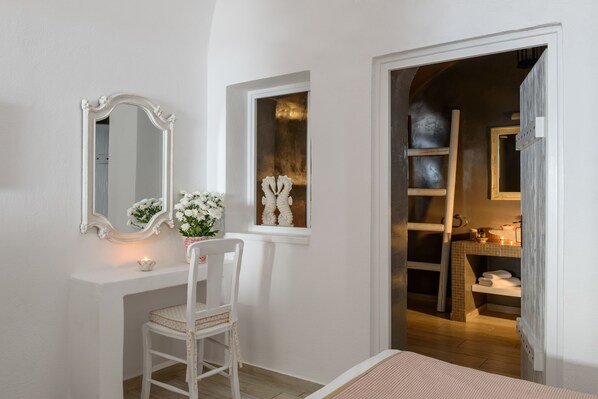 Una habitación con tocador blanco, un espejo, una ventana con un adorno y una puerta que da al baño.