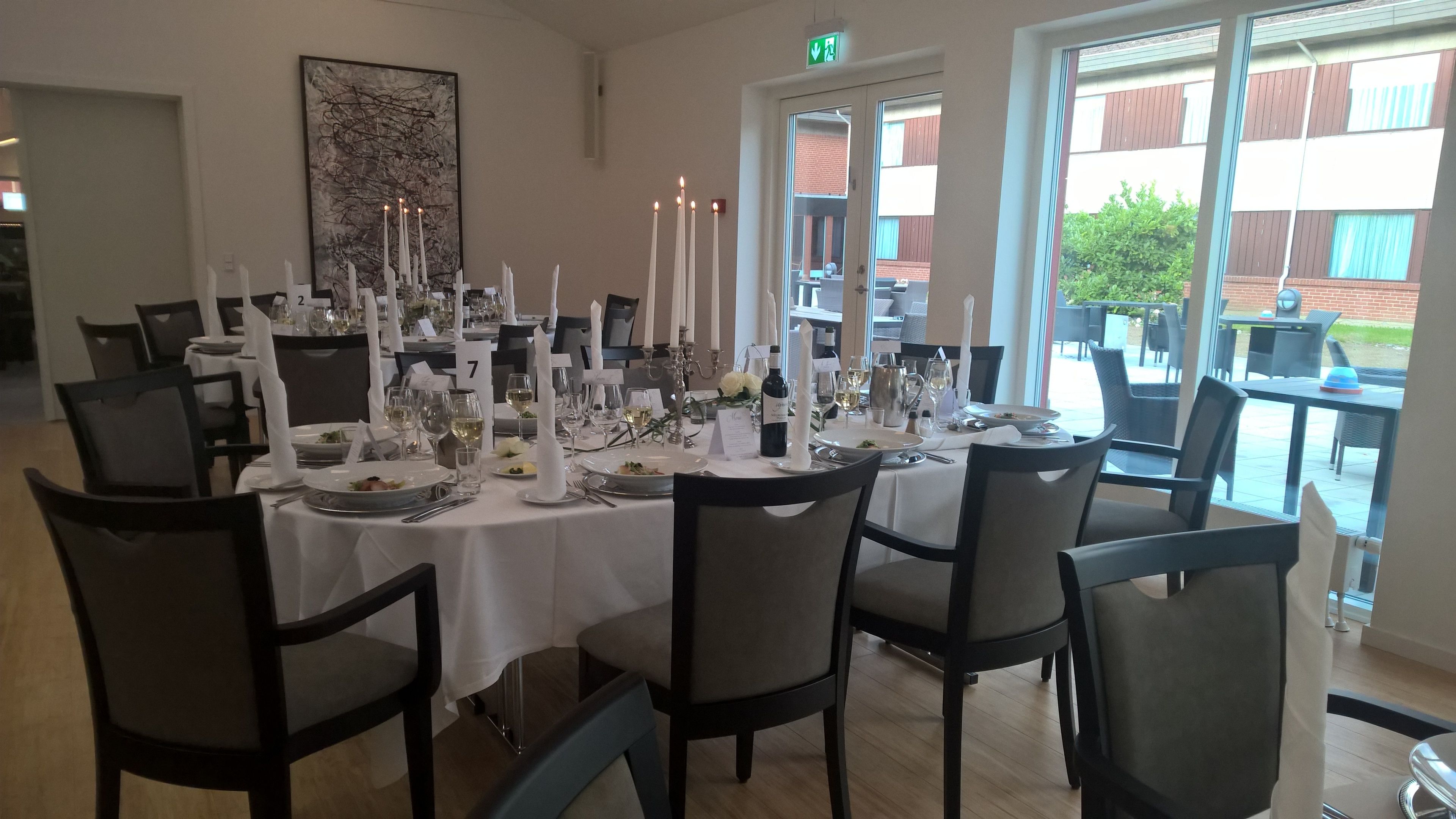 A dining area with tables set for a meal, featuring white tablecloths, candles, and a view of the outside.