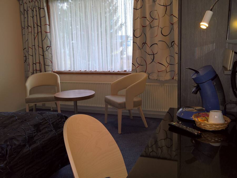 A hotel room with a bed, two chairs, a small round table, a desk with a lamp, a telephone, and a window with curtains.