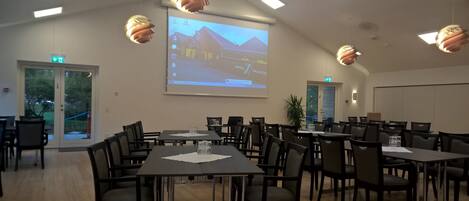 A conference room with multiple tables and chairs, a projector screen displaying a hotel image, and a door leading outside.