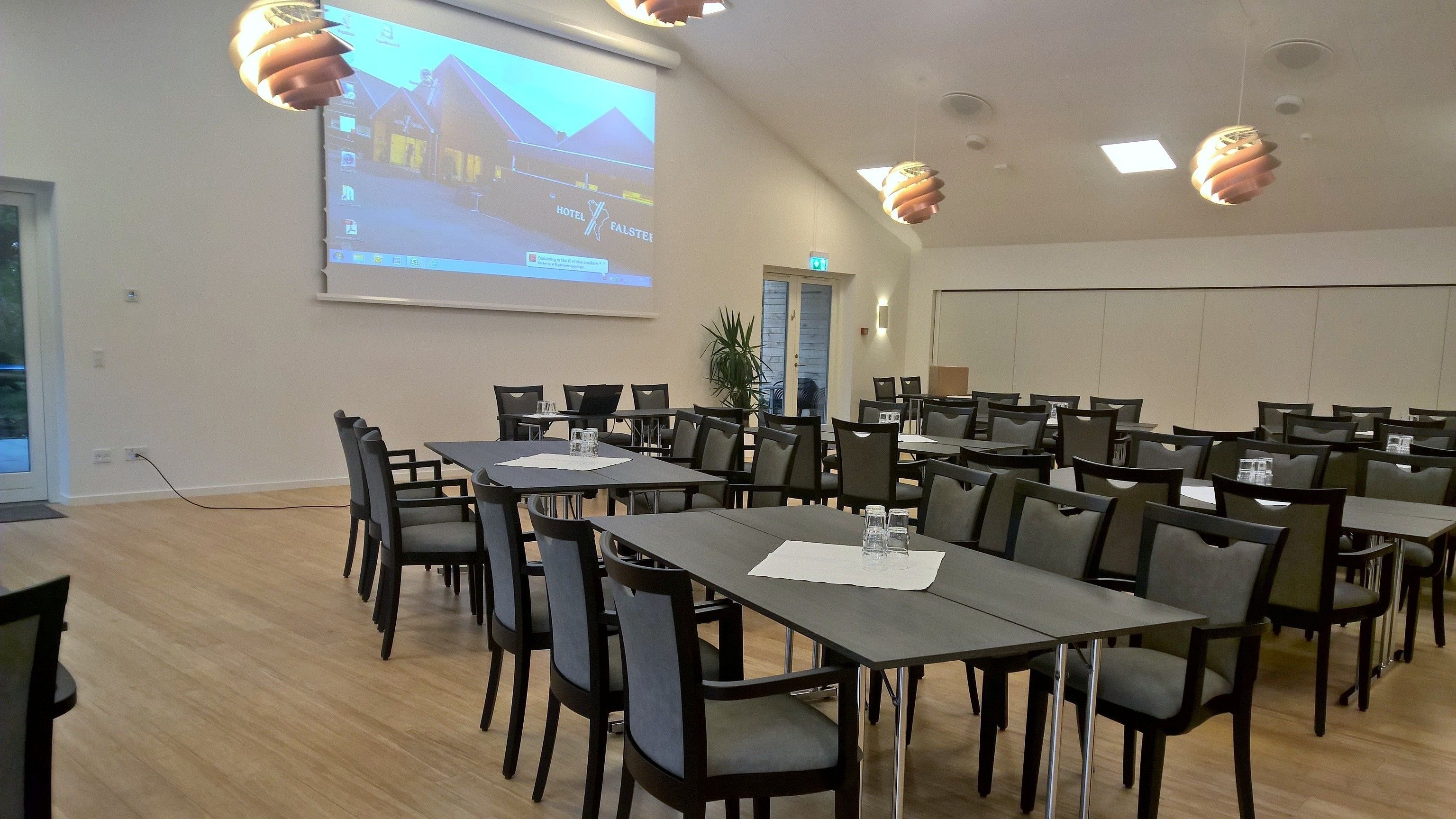 A conference room with multiple tables and chairs, a projector screen displaying a hotel image, and a potted plant.