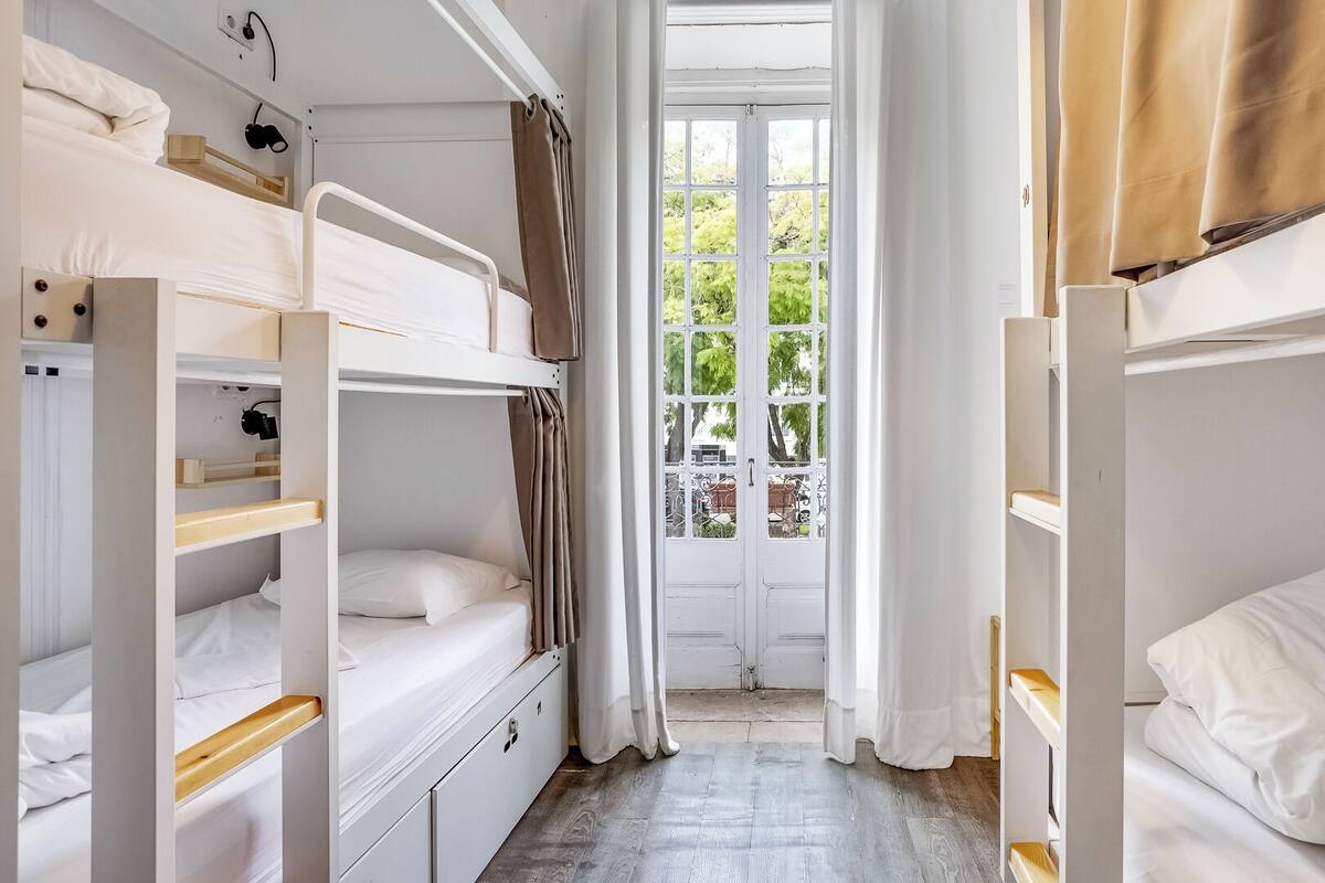 A dormitory room with bunk beds, a window with curtains, and a view of greenery outside.