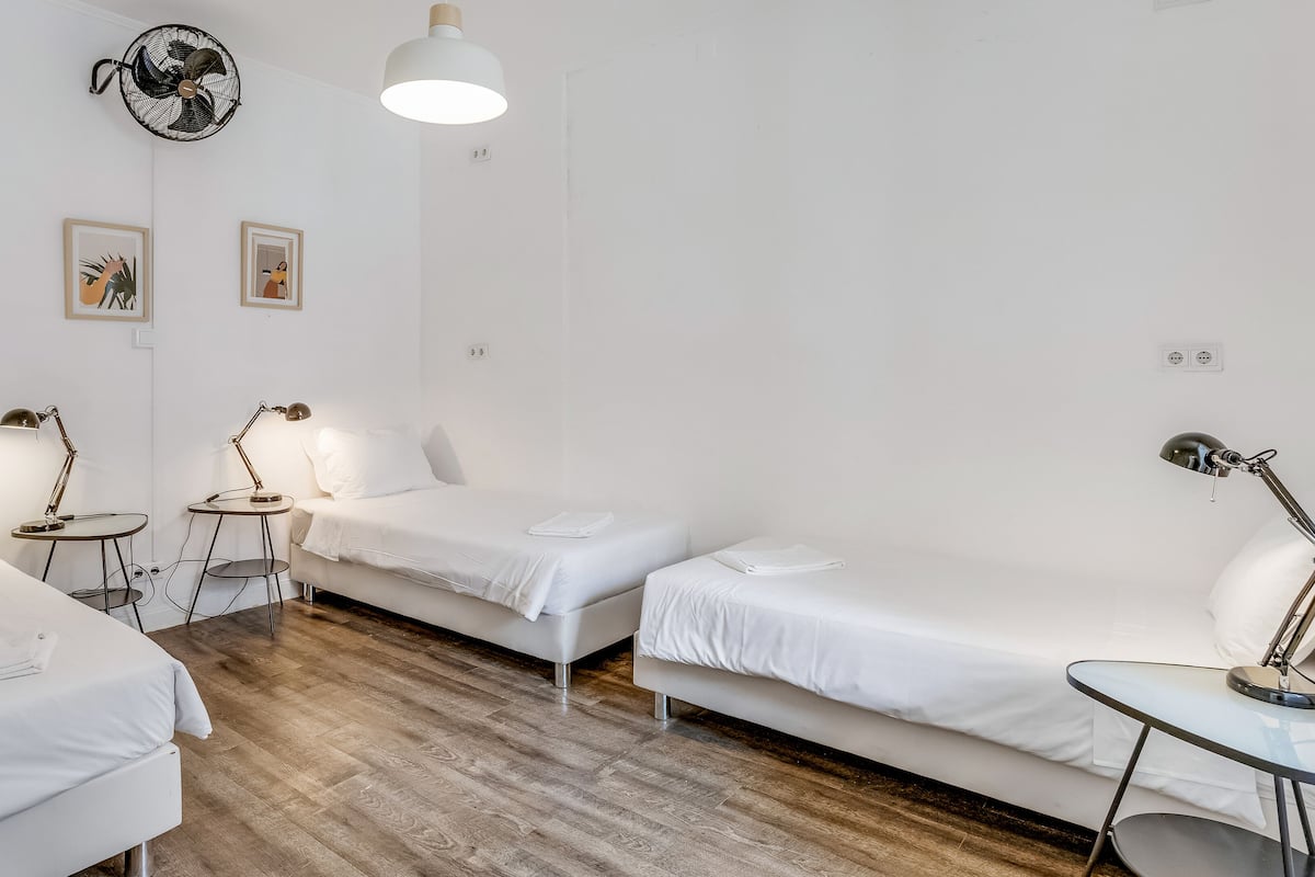 A minimalist hotel room with two single beds, a small table, a standing lamp, and a ceiling fan.