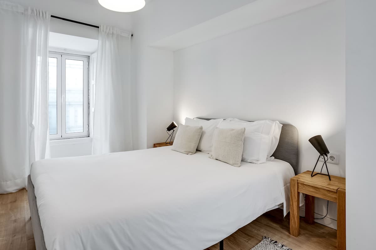 A bedroom with a large bed, white bedding, a wooden nightstand, a floor lamp, and a window with sheer curtains.