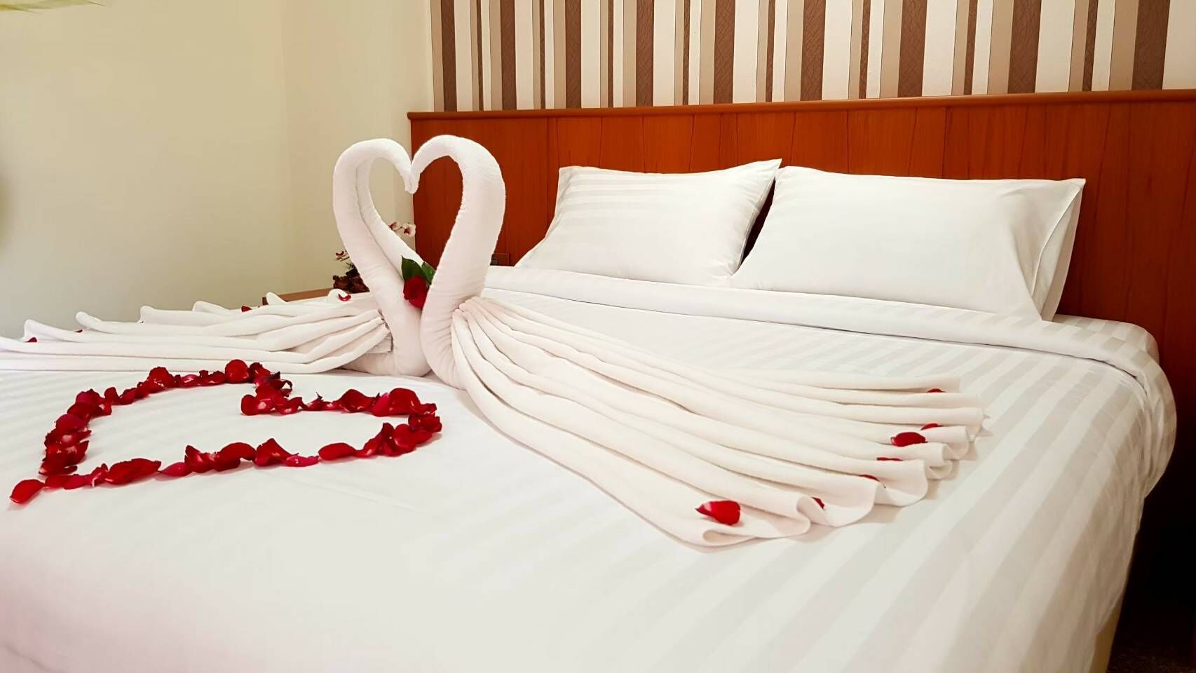 Un lit parfaitement fait, avec du linge de lit blanc, une composition en forme de cœur faite de pétales de roses et deux décorations en forme de cygne, réalisées à partir de serviettes.