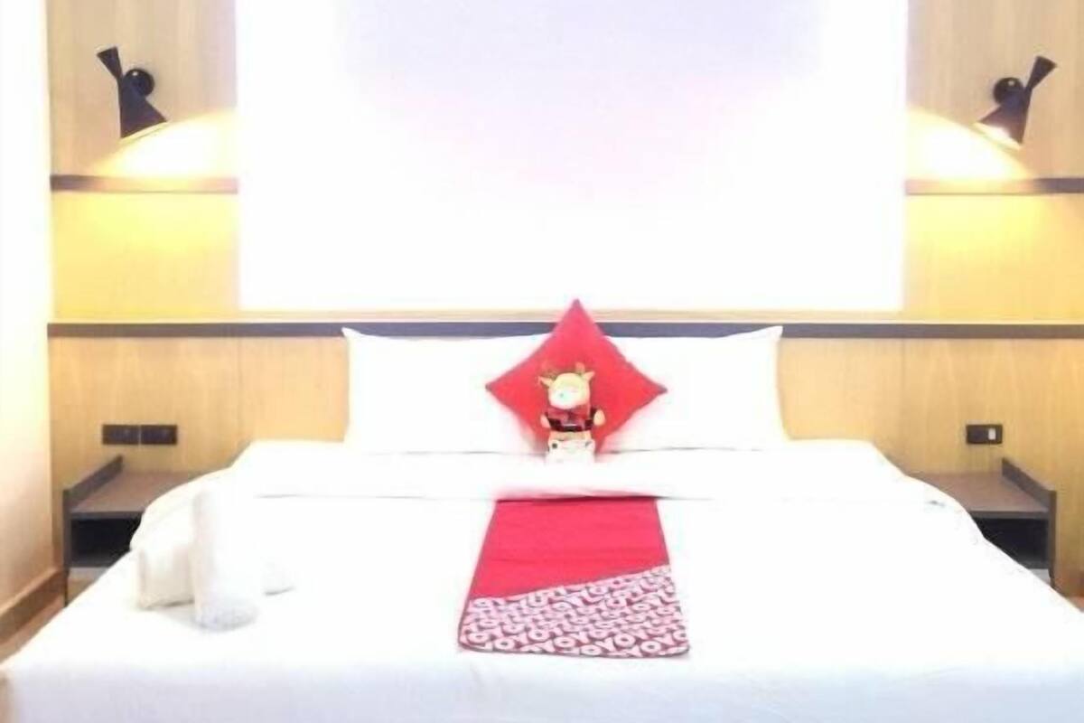A hotel room with a bed, a red pillow, a white pillow, a red and white throw, and two wall-mounted lamps.