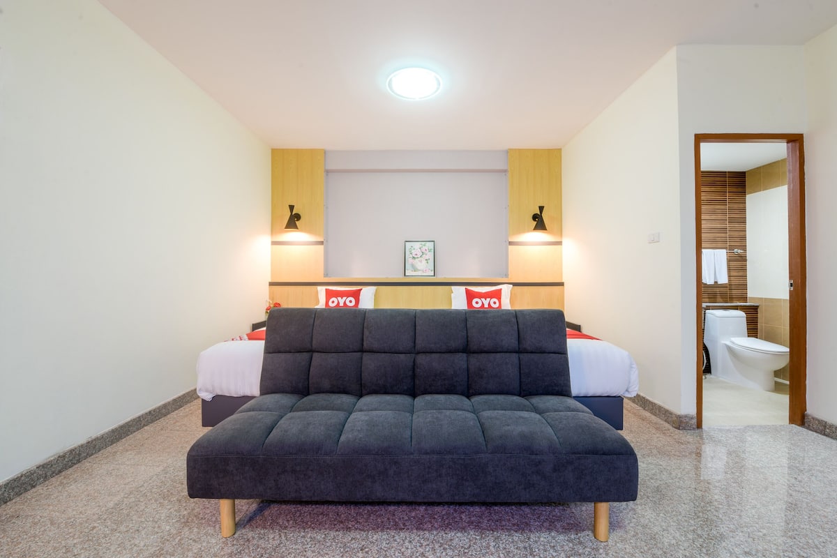 A modern hotel room with a large dark sofa, two beds, a wooden headboard, and a bathroom visible through an open door.