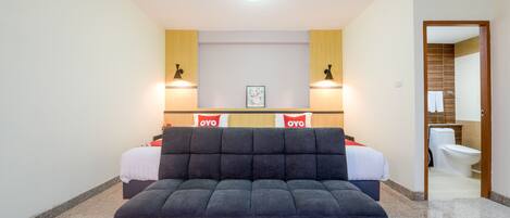 Una habitación de hotel moderna con un sofá grande en tonos oscuros, dos camas, un cabecero de madera y un baño visible a través de una puerta abierta.