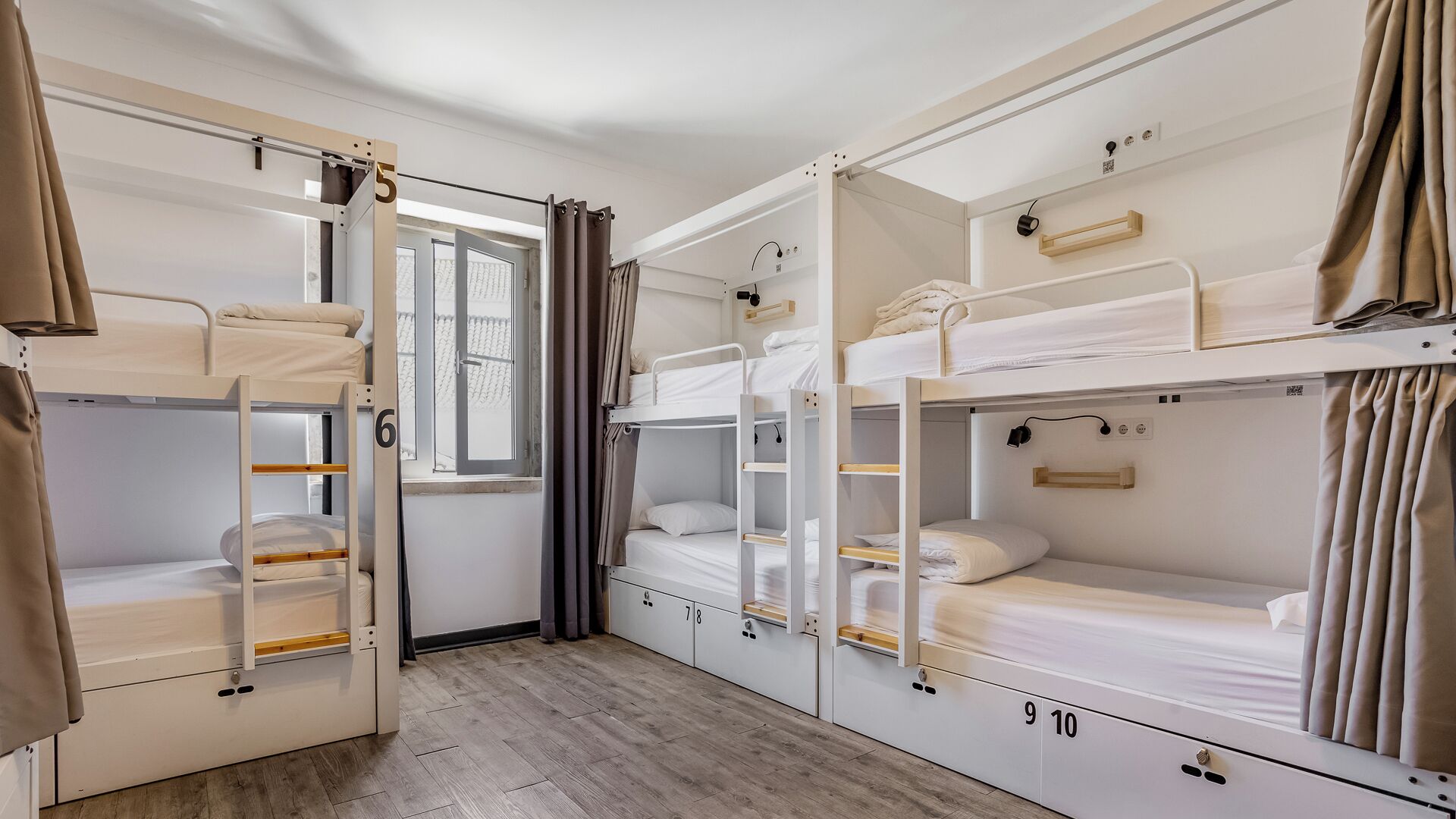 A dormitory room with bunk beds, numbered storage compartments, and a window with curtains.