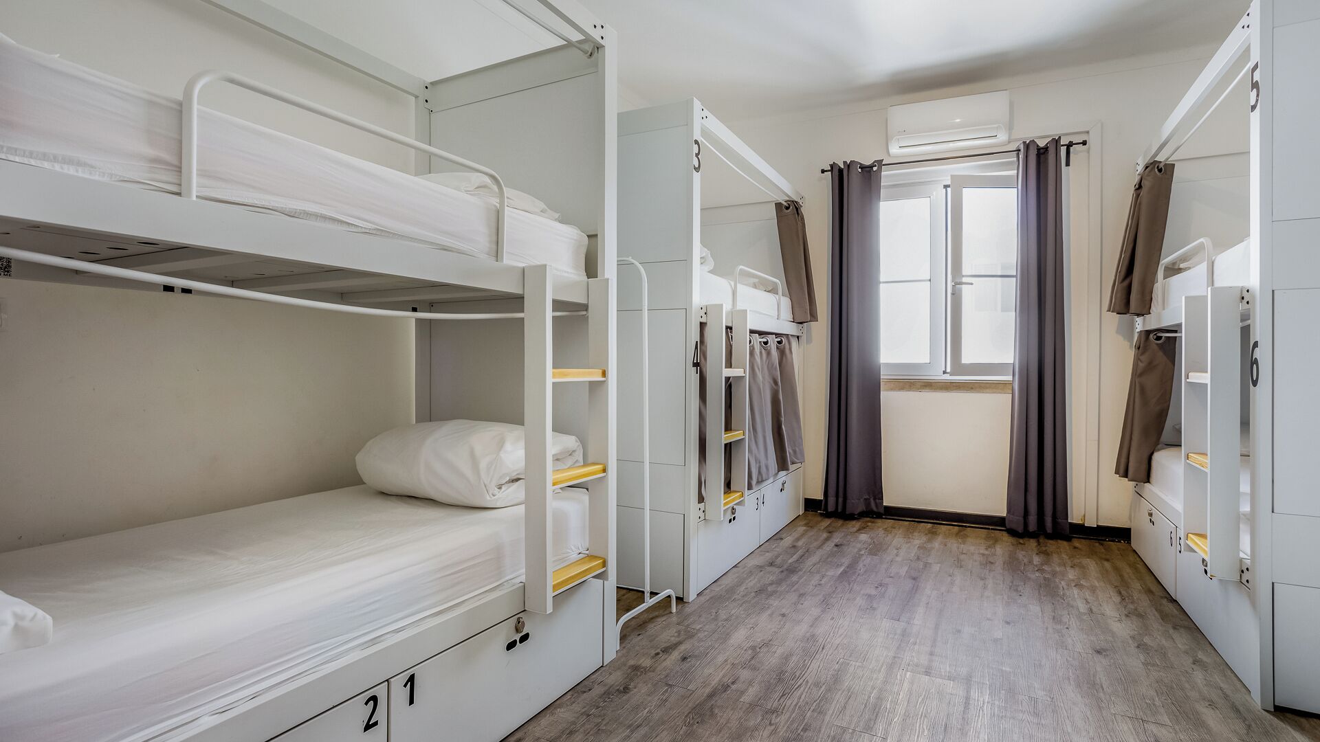 A dormitory room with bunk beds, a window with curtains, and a wooden floor.
