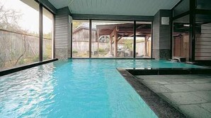 Piscina interior com paredes de vidro transparente, borda de pedra e vista para um deck de madeira e uma casa ao fundo.
