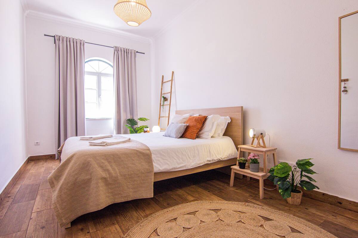 Une chambre avec un plancher en bois, un lit doté d’une tête de lit blanche, une table de chevet avec une lampe et une fenêtre avec des rideaux.
